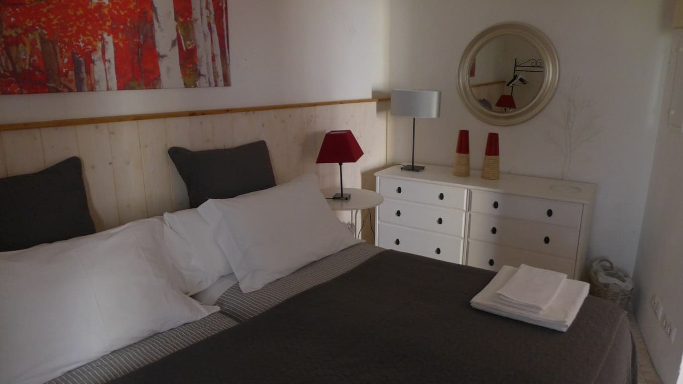 Camas twin com colcha cinzenta ao lado de uma cómoda branca, sob um espelho redondo e um quadro vermelho