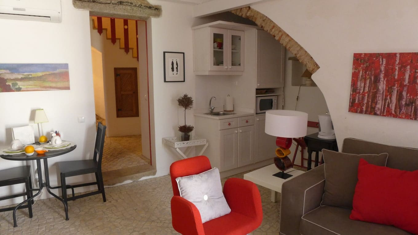 Sala com um cadeirão vermelho sob um arco de pedra, com a kitchenette e a mesa de refeições ao fundo