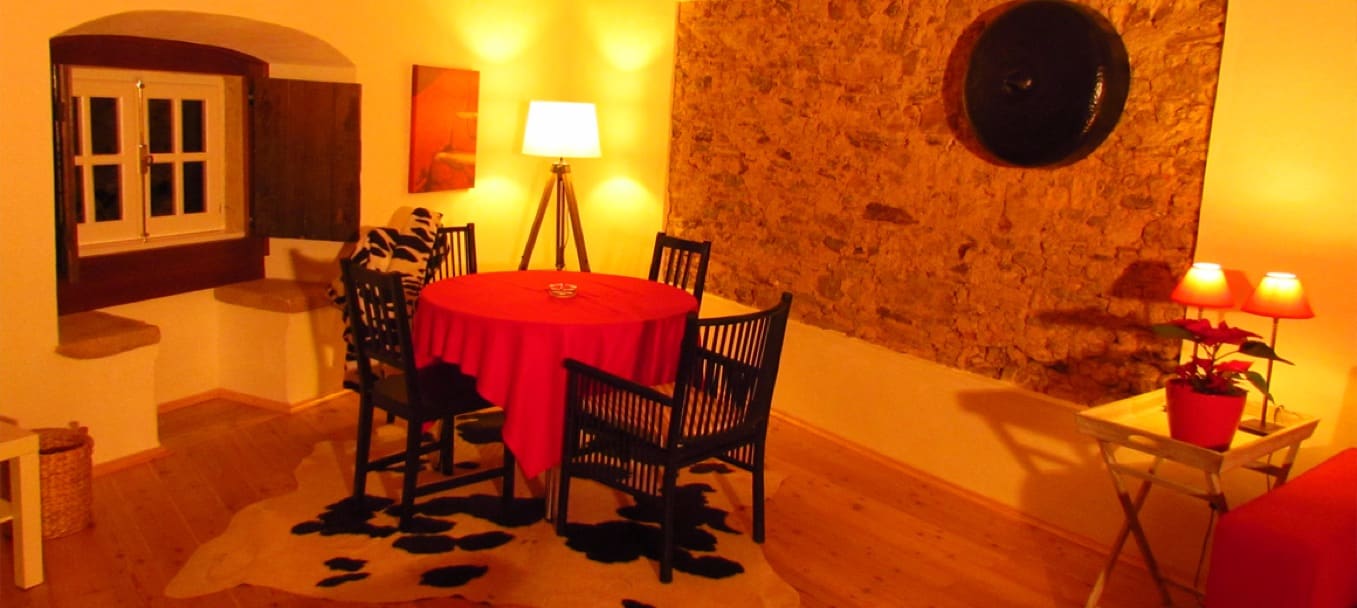 Mesa redonda posta com toalha vermelha junto a uma parede de pedra, iluminada por candeeiros ao fim do dia